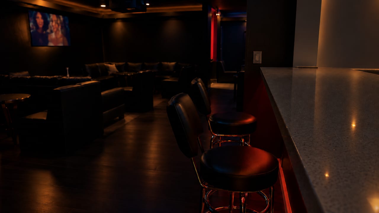 Two black leather bar stools at a stone-topped bar counter, with leather lounge seating and a wall-mounted screen in the dim room beyond.