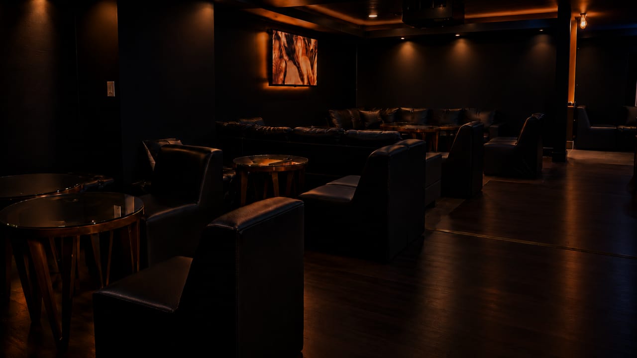 A deep view across the lounge: leather armchairs and sofas, round glass-topped tables, and a backlit screen on the far wall.