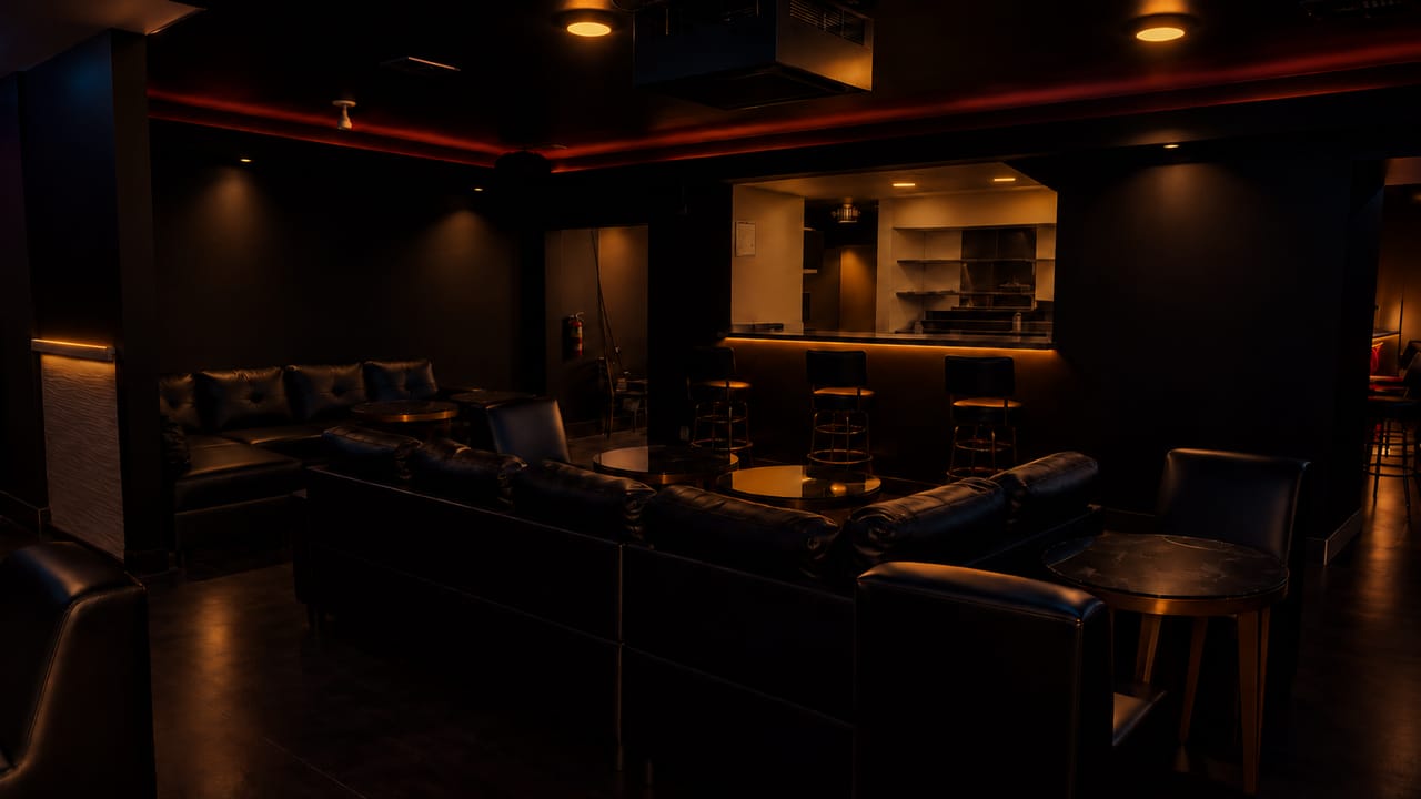 Leather lounge seating facing the bar, where three bar stools line a lit counter opening beneath red and amber ceiling light.