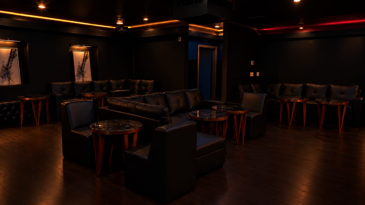 Wide view of the upstairs lounge: black leather sofas and armchairs around round copper-legged tables, with amber and red light along the ceiling.