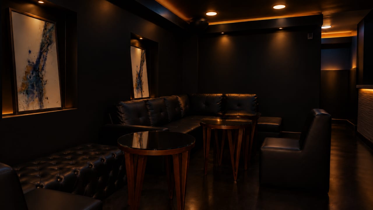 An intimate lounge room with tufted black leather seating, round glass-topped tables, and two abstract paintings lit in recessed wall niches.