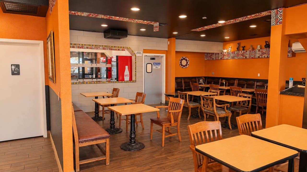 Wide view of the main dining room: orange walls, woven geometric trim along a dark ceiling, wooden chairs and rows of tables beneath recessed lights.